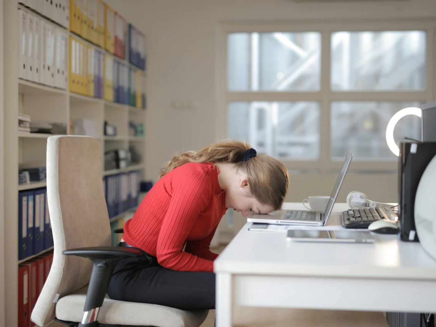 Woman in a red sweater, sitting at a desk with her head resting on a laptop, surrounded by shelves of binders.