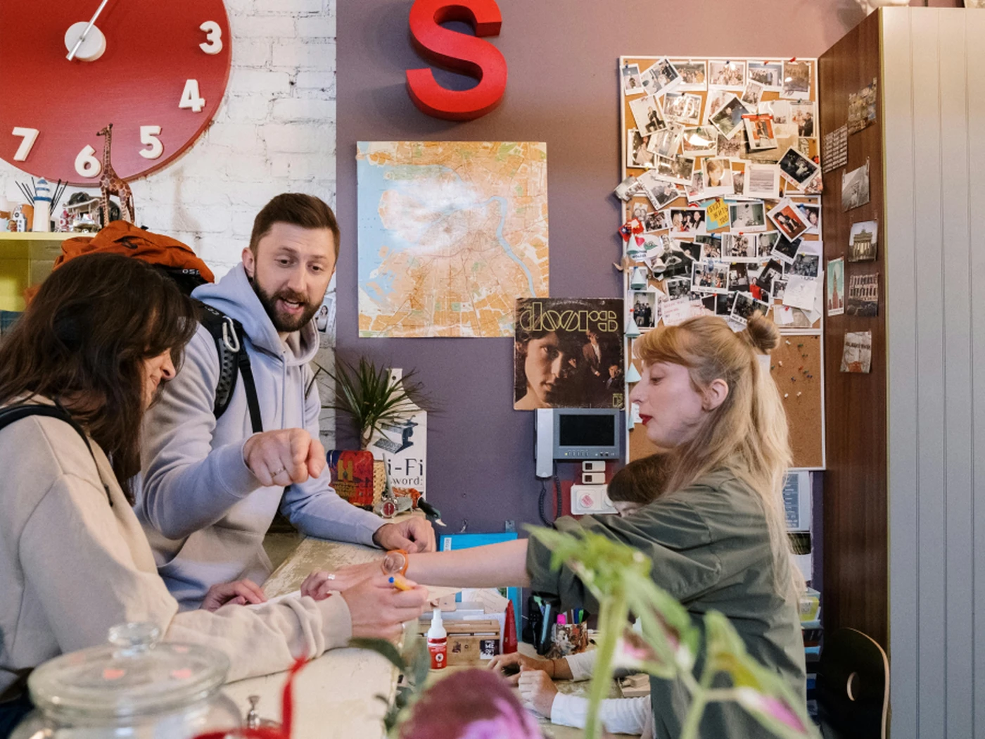 People checking in at a hotel front desk counter with a colorful wall, map, and photos. A large red clock and letter "S" are visible in the background.