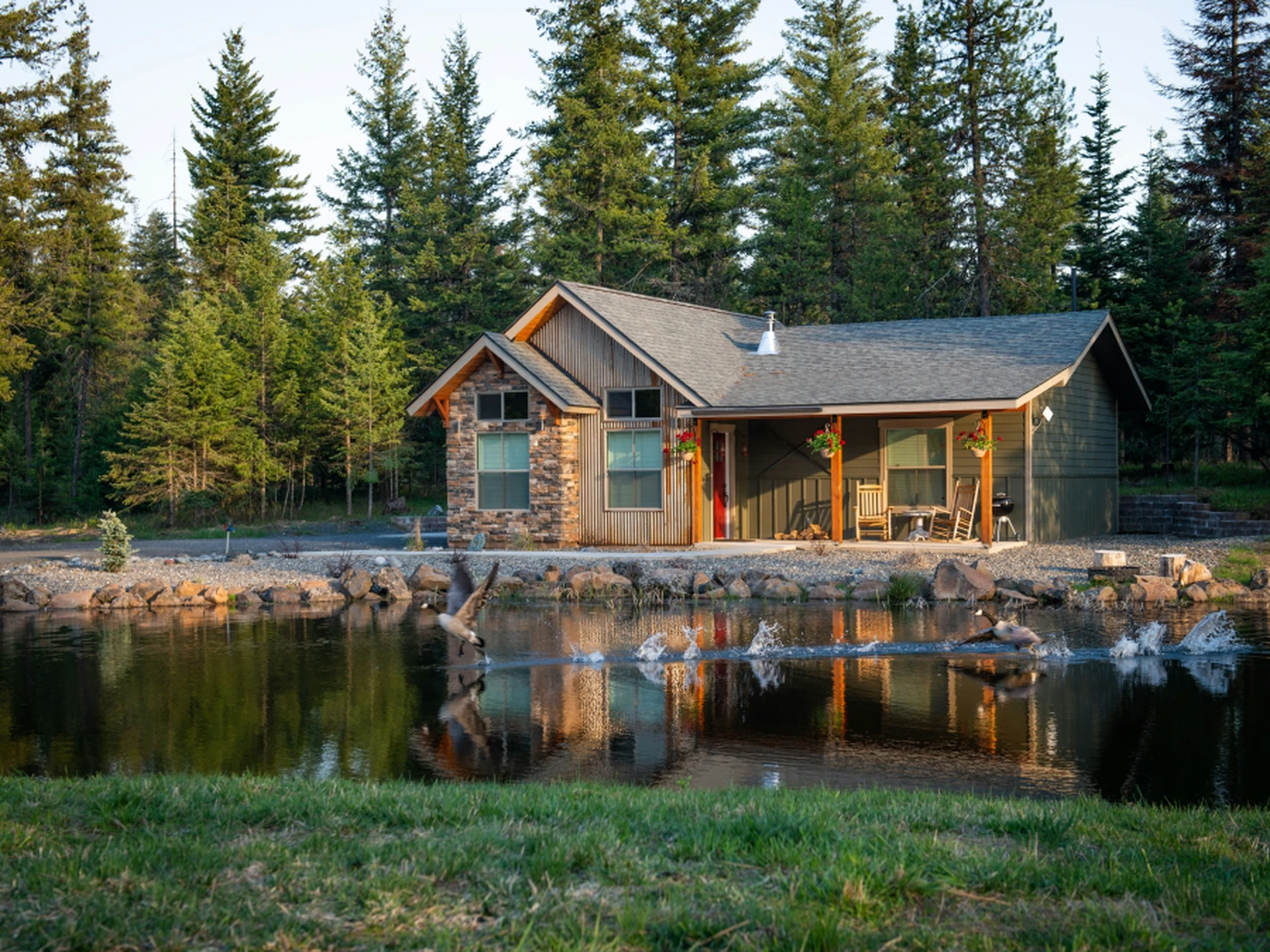 A cozy cabin with a stone facade by a tranquil pond, surrounded by tall pine trees under a clear blue sky.