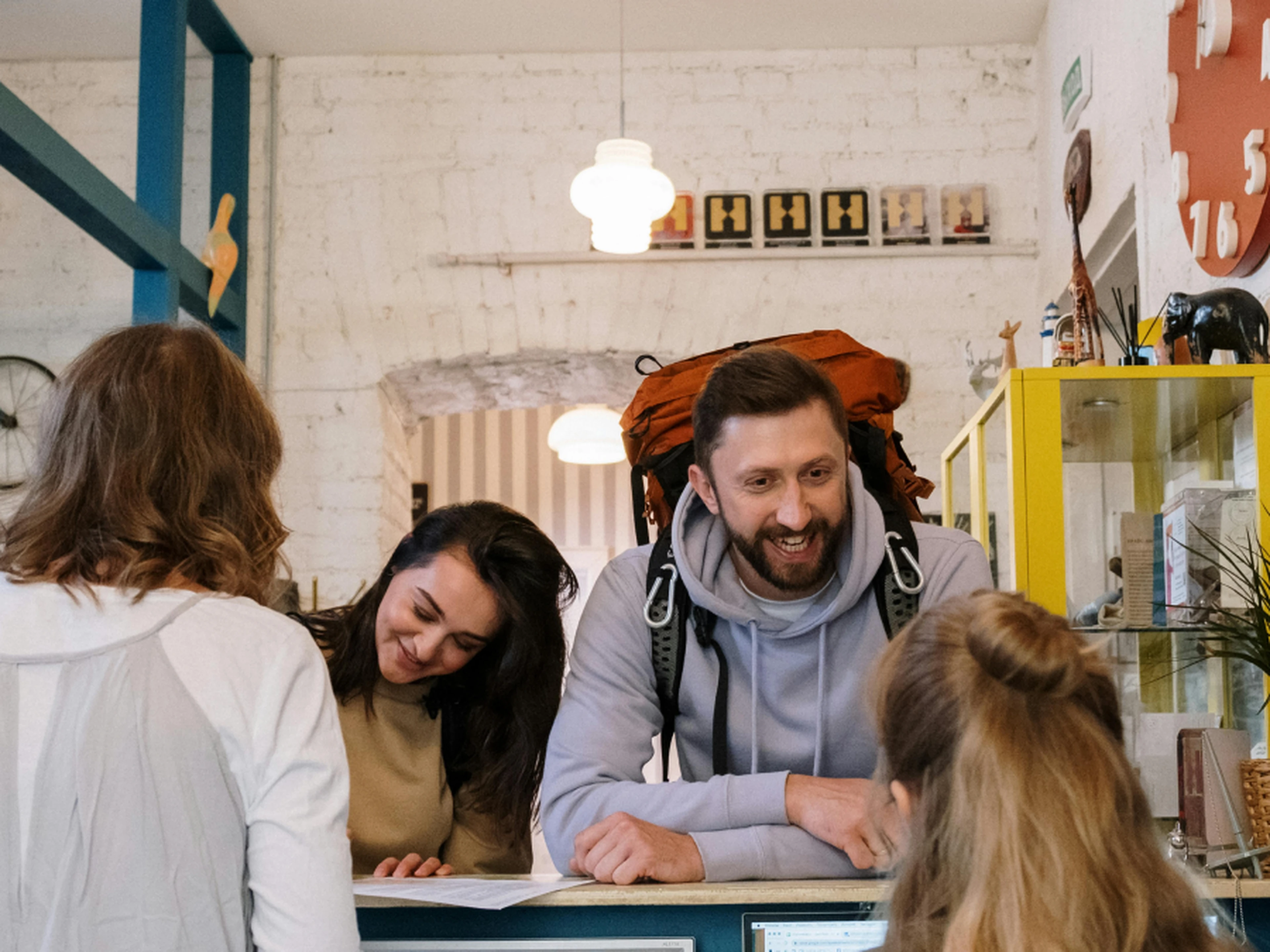 People at a reception desk in a cozy setting, with one person wearing a backpack and engaging in conversation with the staff.
