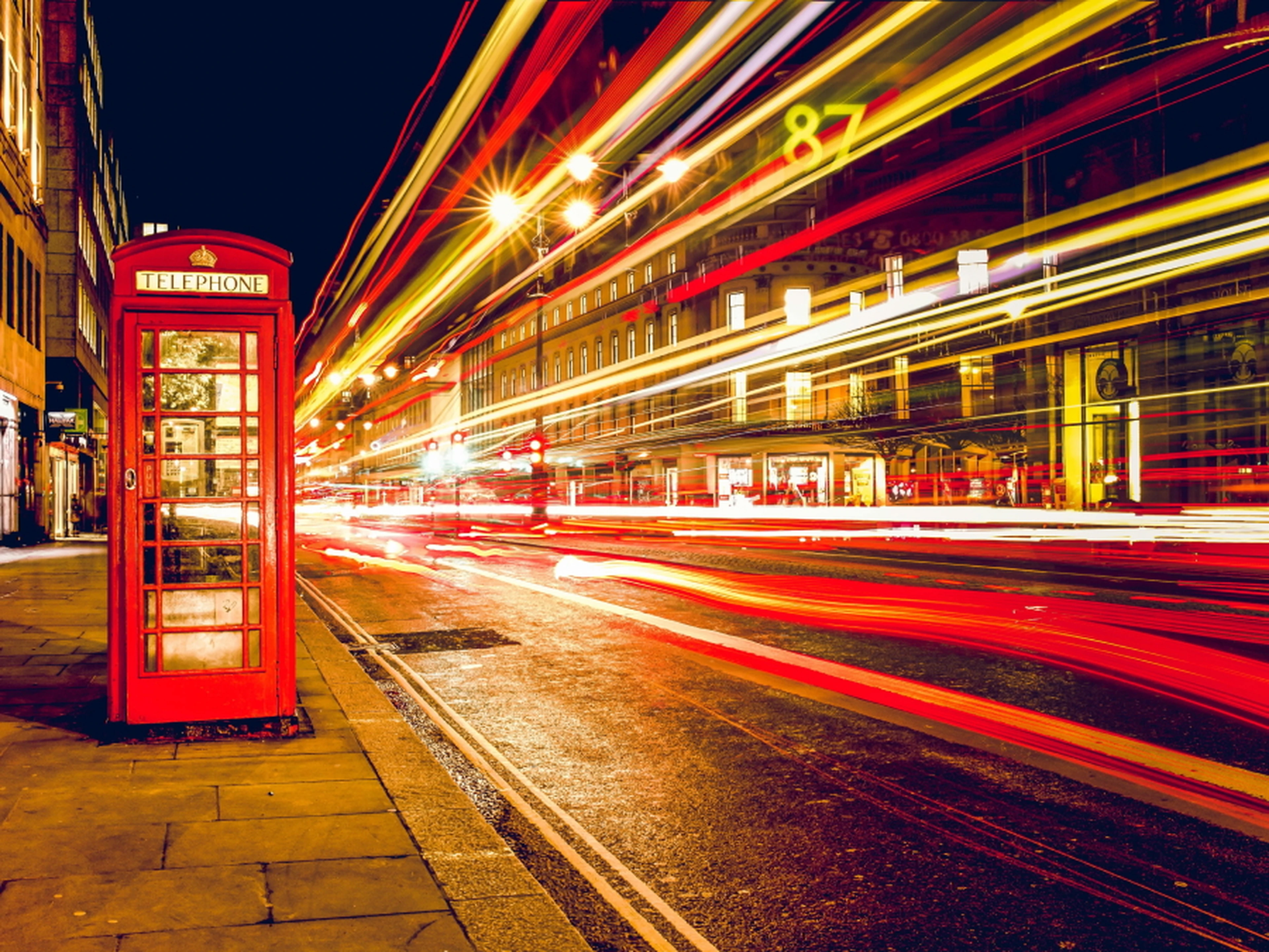 A red telephone booth stands on a city street at night, with vibrant light trails from passing vehicles creating dynamic streaks.