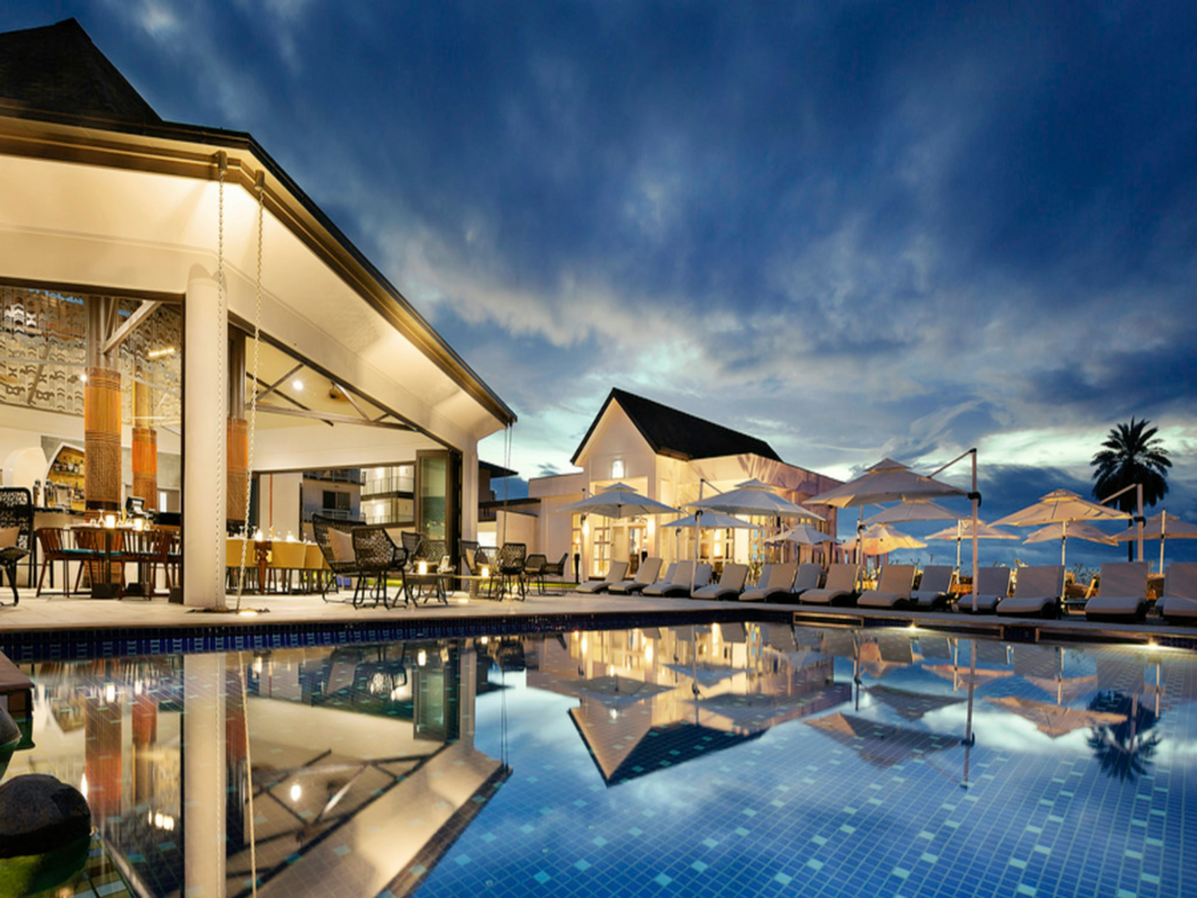 Luxurious resort poolside at dusk, with elegant lounge chairs, umbrellas, and a modern open-air dining area reflecting in the water.