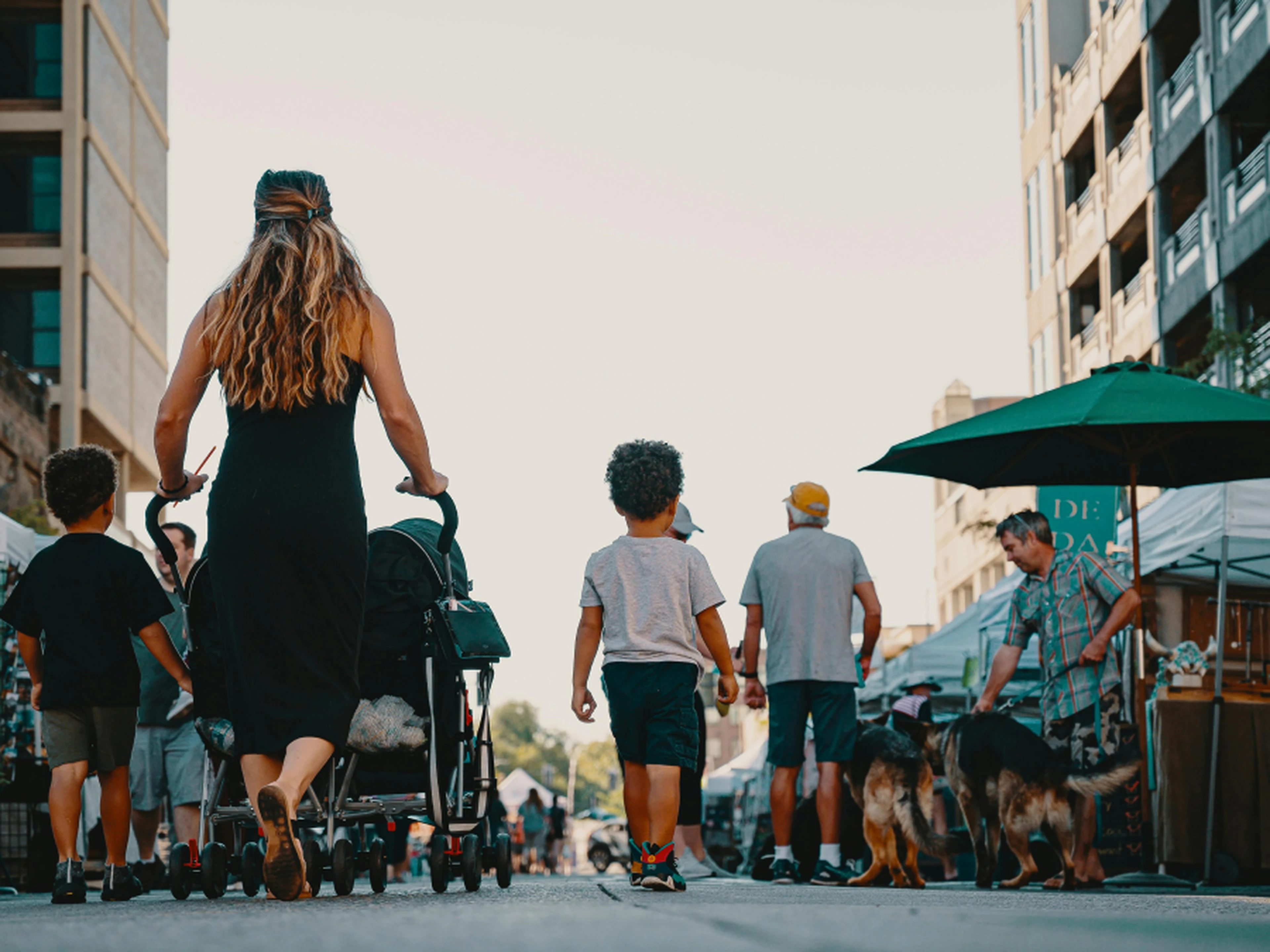 A woman pushes a stroller down a bustling street market, surrounded by people and dogs, with tall buildings lining the background.