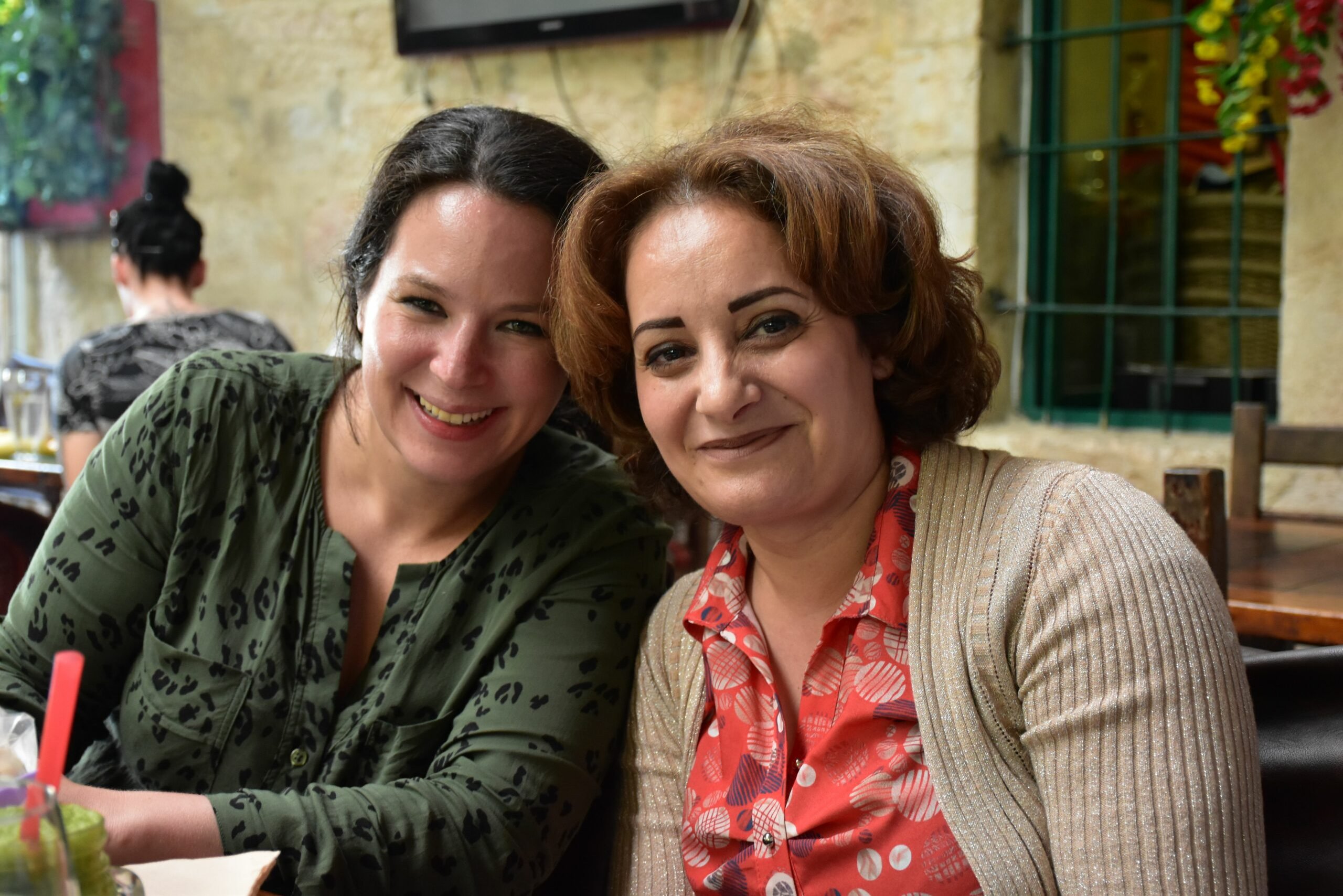 Enaam and Helena smiling at a table in a cozy café, with a stone wall and window in the background.