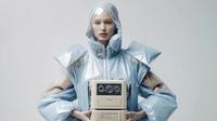 Person in futuristic, angular blue outfit holds a vintage tape recorder against a neutral background.