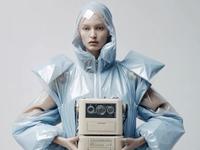 Person in futuristic, angular blue outfit holds a vintage tape recorder against a neutral background.