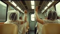 Two people in plastic ponchos raise their arms inside a train carriage, with other passengers seated nearby.