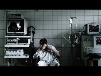 Person in a hospital gown sits in a medical room, surrounded by equipment and monitors, with a distressed expression.