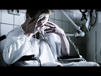 Person in a hospital gown sitting in a wheelchair, holding their head with wired medical sensors attached, in a clinical setting.