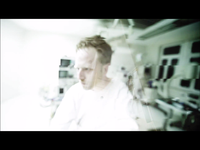 Blurred image of a person in a white room with medical equipment, creating a distorted, abstract effect.