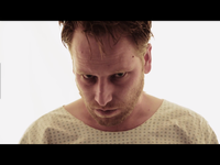 Man with disheveled hair and intense expression wearing a hospital gown, looking downward against a bright background.