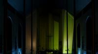 A dimly lit church interior with colored lighting, highlighting a cross and altar at the center against tall walls and arched windows. Projection over the altar.