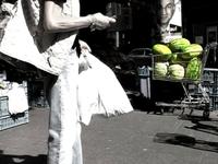 Low saturated Image showing a person holding bags and phone, walking past a shopping cart filled with green watermelons on a sunny street.