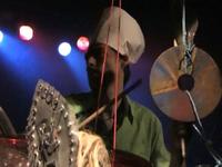 Musician playing drums and cymbals on stage, wearing a white chef's hat, with colorful stage lights in the background.