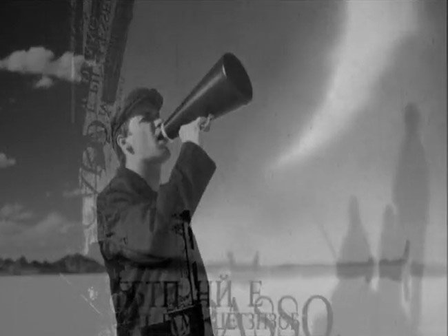 Black and white image of a young man speaking in a megaphone in front of a fluffy cloud sky.
