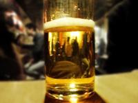 A glass of beer on a table reflects a blurred, dimly lit bar scene with people in the background.