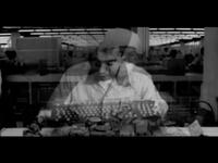 A black and white image of a person working on electronics, with a blurred, double exposure effect in a factory setting.