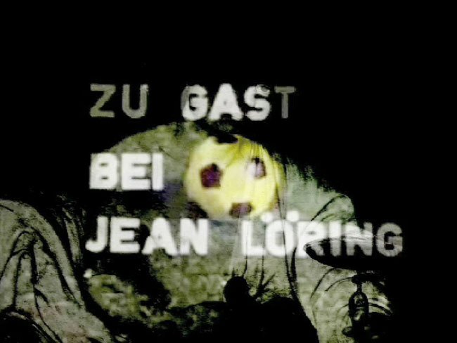 The image shows the text "Zu Gast bei Jean Löring" with a soccer ball graphic, projected onto a dark background.