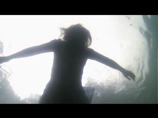 Silhouette of a person floating underwater with arms outstretched, viewed from below against a bright surface.