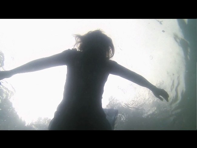 Silhouette of a person floating underwater with arms outstretched, viewed from below against a bright surface.