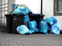 Low saturated image of blue garbage bags piled on and around black bins in an outdoor urban setting.