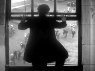 Black and white still image. A person in silhouette stands at an open window, looking down at a street filled with people below.