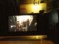 Camera screen showing a dimly lit bar scene with people gathered, surrounded by the camera body in the foreground.