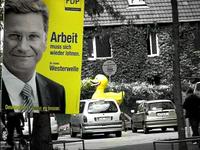 Low saturated image of political poster from an unimportant party with a man's face and German text beside a street with parked cars and a large yellow duck statue in the background.