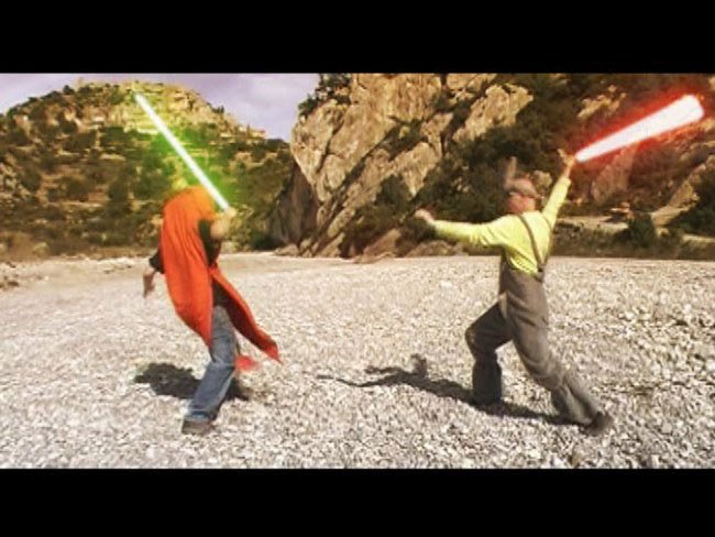 Two people, one in carrot and one in a rabbit costume, in a rocky landscape are engaged in a lightsaber duel, one with a green blade and the other with a red blade.