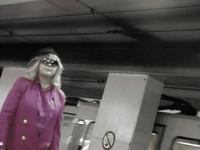 Low saturated image of a person in a bright pink coat and sunglasses stands on a subway platform, with a train in the background.