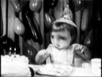 Black and white photo of a child wearing a party hat, sitting at a table with a cake. Balloons are in the background.
