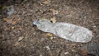 A crushed plastic bottle lies on a forest floor covered in dry leaves and twigs.