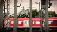 Red train passing behind a statue of a horse and rider, framed by metal poles and power lines, under a cloudy sky.