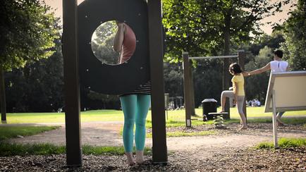 Person standing behind a circular playground structure, partially hidden. Other people are in the background, enjoying the park.