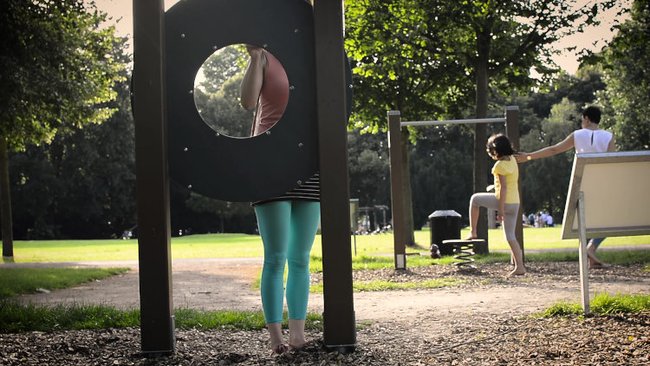 Person standing behind a circular playground structure, partially hidden. Other people are in the background, enjoying the park.