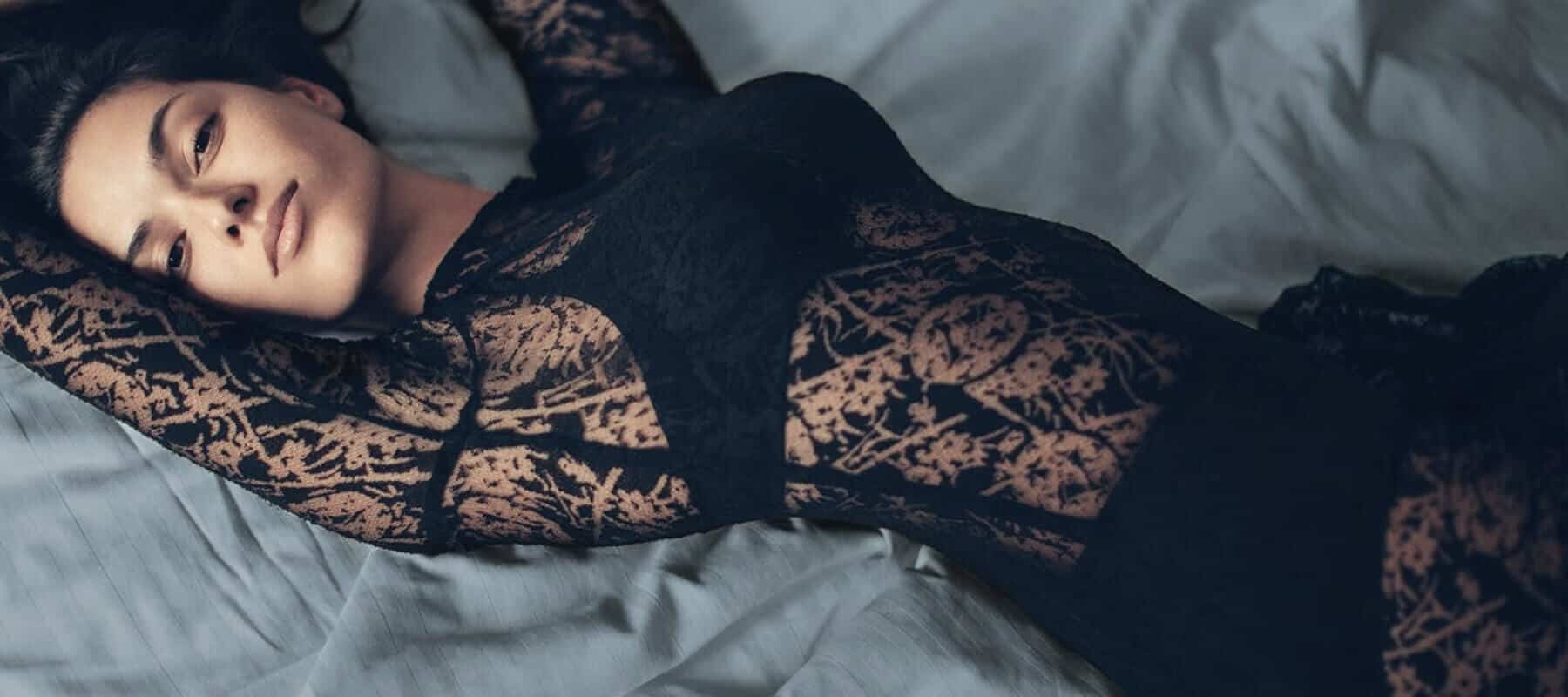 Person lying on a bed, wearing a black lace dress with intricate patterns, arms above head, looking relaxed and serene.