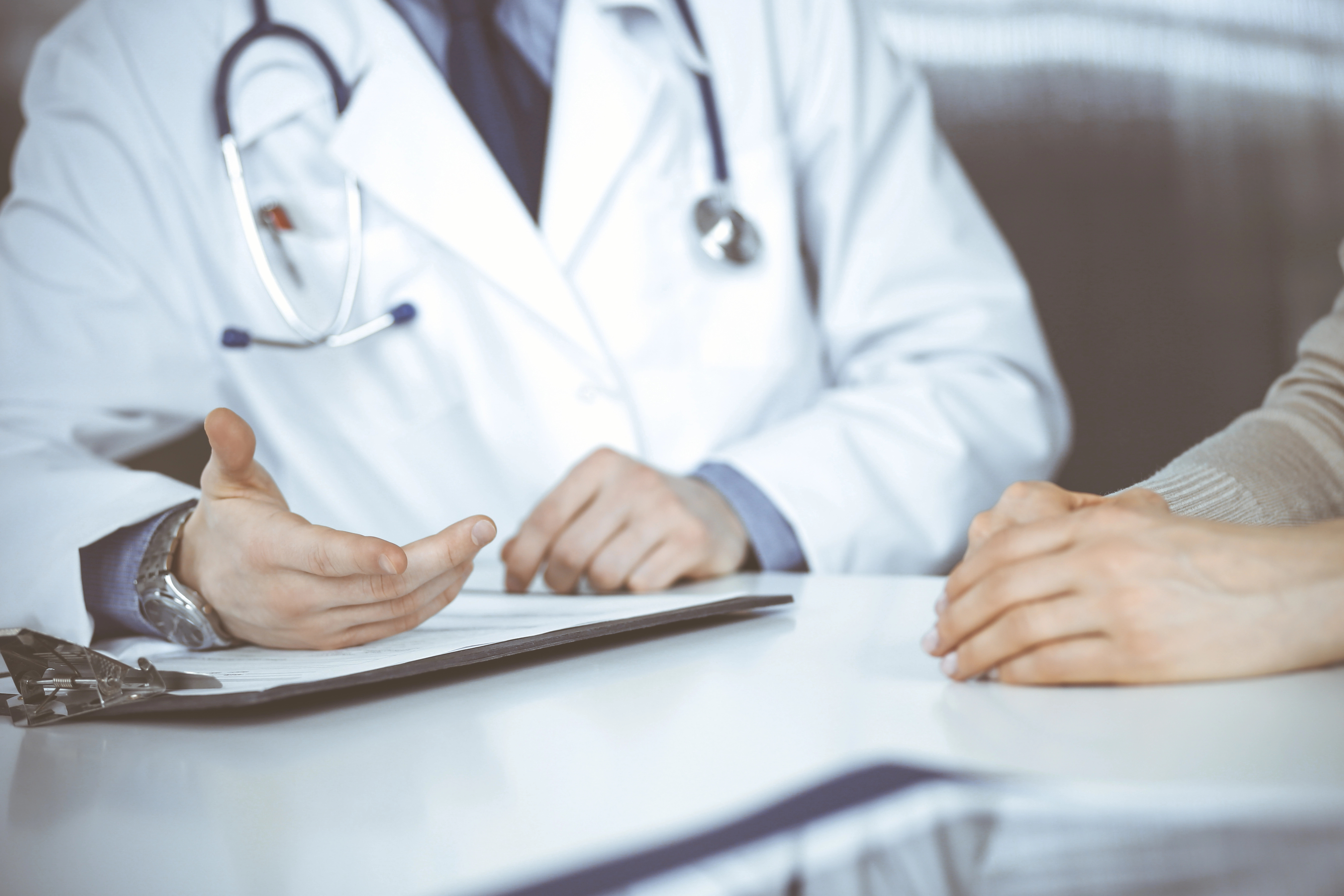 Doctor in a white coat discussing with a patient across a desk, using hand gestures to explain.