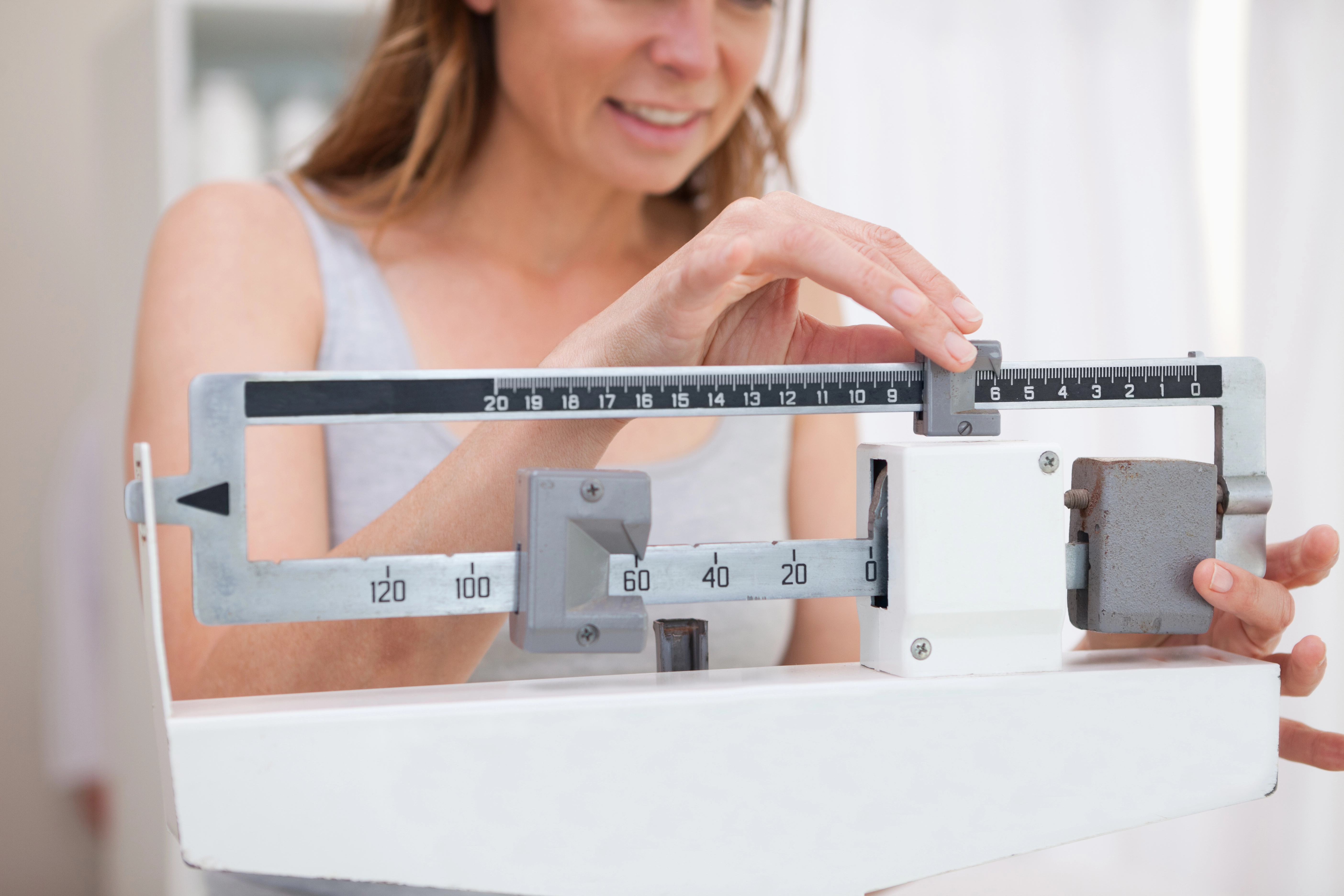 Woman adjusting the weights on a balance scale, smiling as she checks her weight in a bright room.