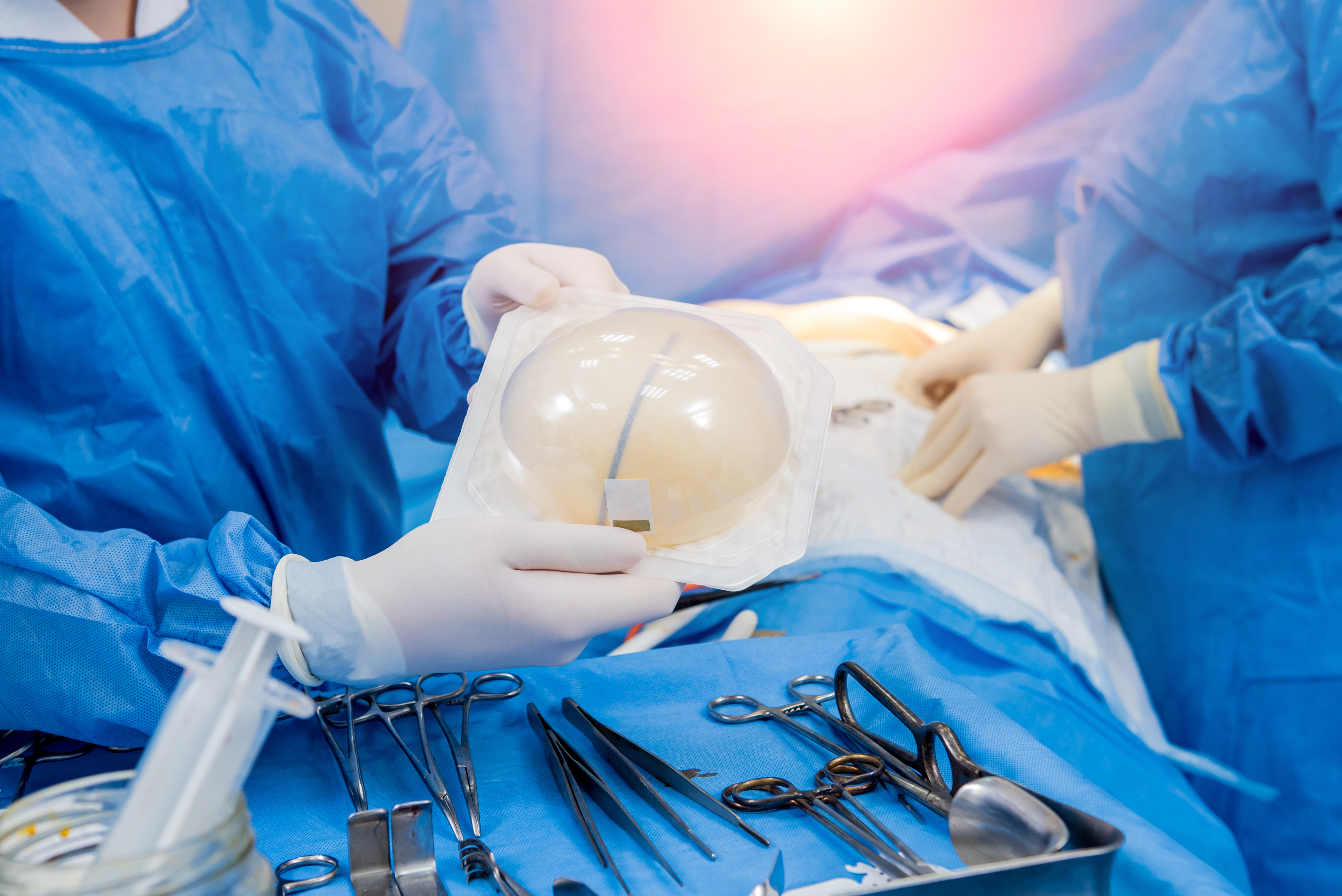 Surgeons in blue gowns and gloves hold a breast implant in an operating room, surrounded by surgical instruments on a tray.