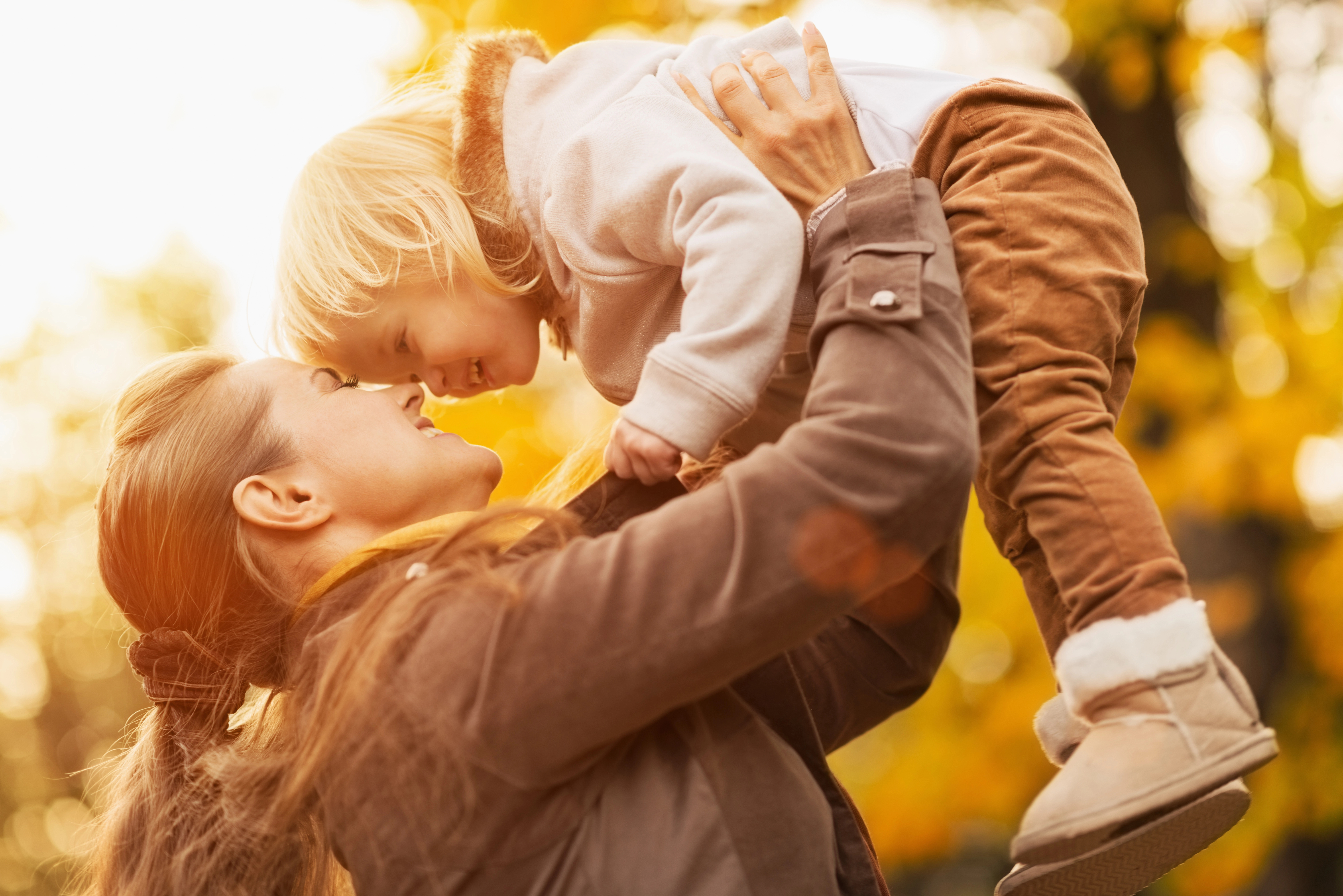 A woman lifts a smiling child in the air during autumn, with warm light filtering through colorful foliage in the background.