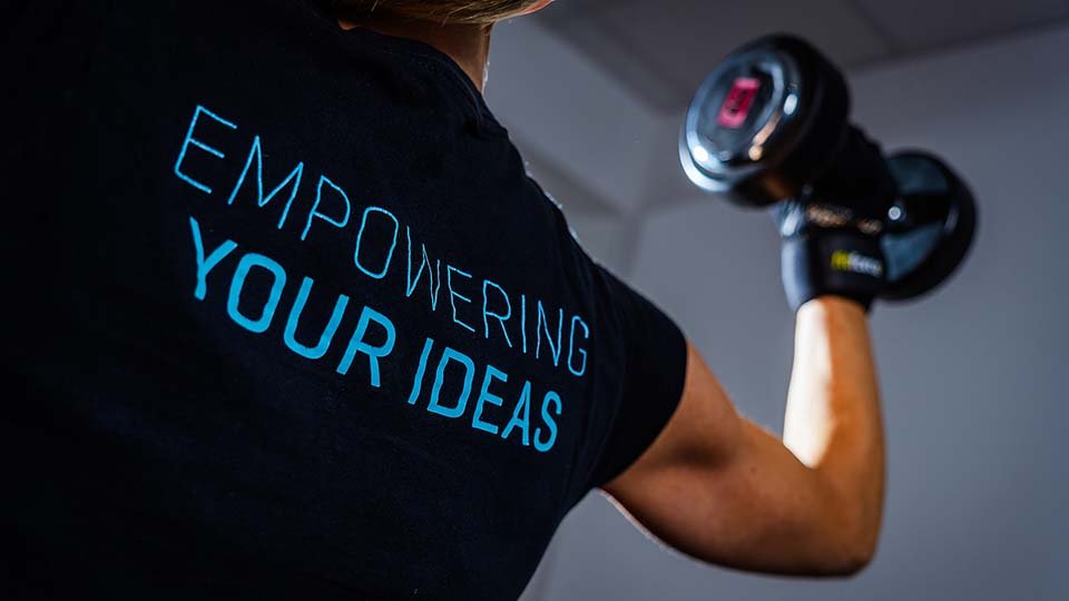 Person lifting a dumbbell while wearing a shirt with "Empowering Your Ideas" printed on the back, in a brightly lit room.