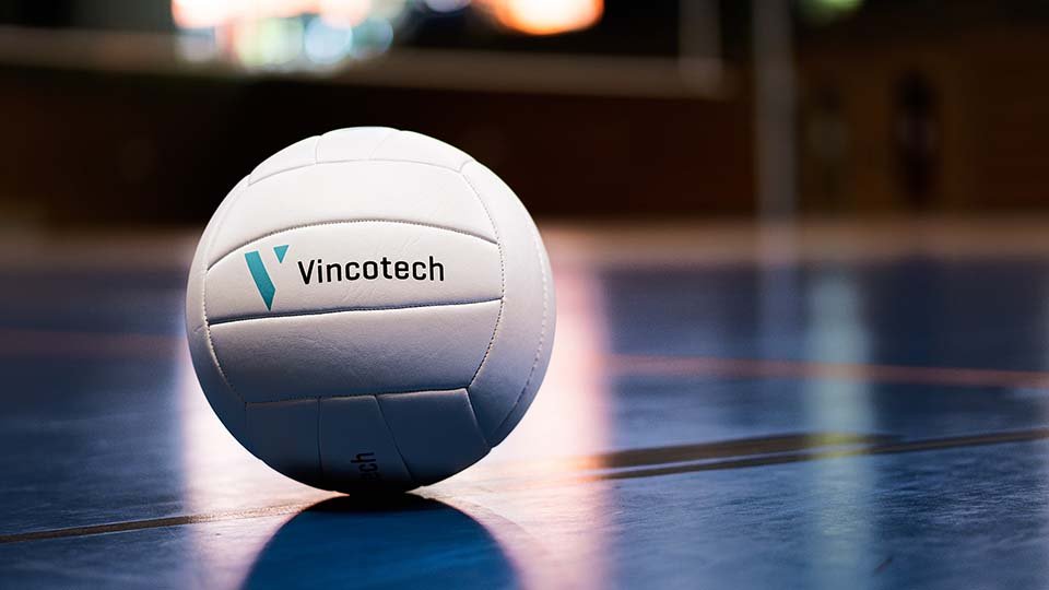 A white volleyball with "Vincotech" logo sits on a polished indoor court, with blurred background.