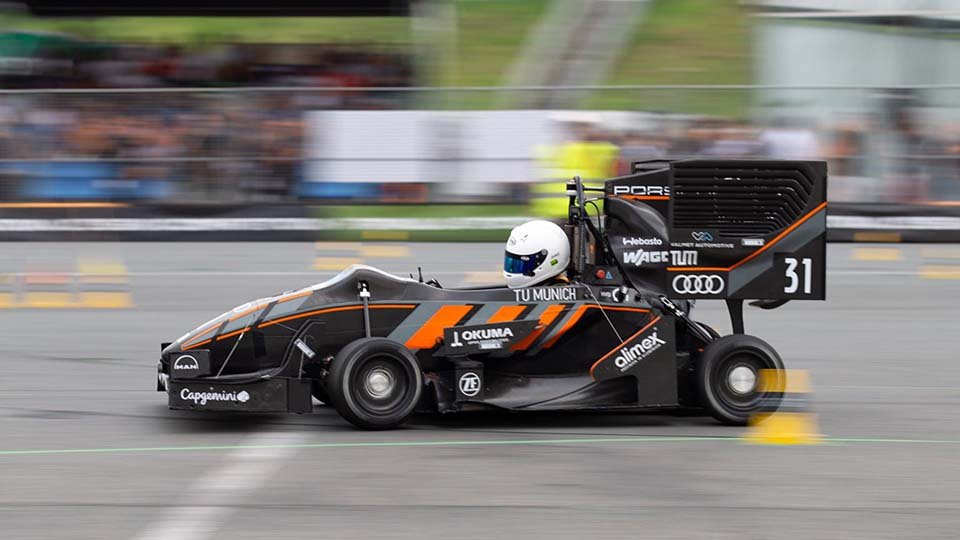 Driver in a black and orange Formula Student race car with visible sponsors, speeding on a track, blurred background, helmeted driver focused ahead.