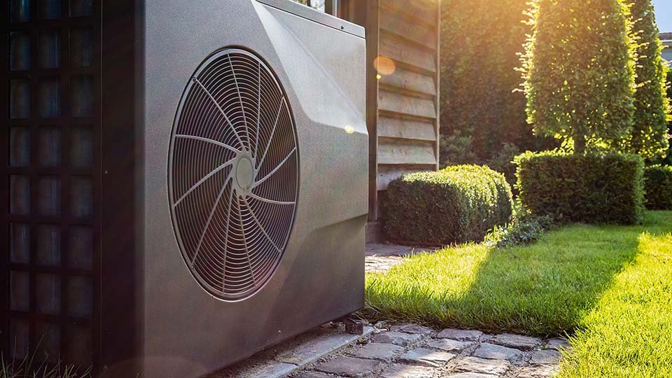 Outdoor heat pump unit next to a house, surrounded by neatly trimmed hedges and a sunny garden path.