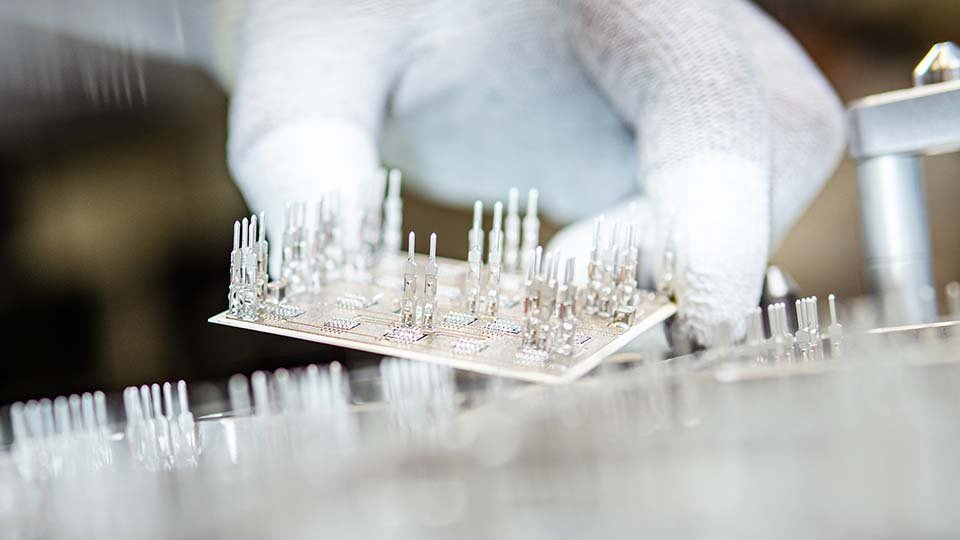 Close-up of a glove holding a PCB with press-fit pins in a production setting.