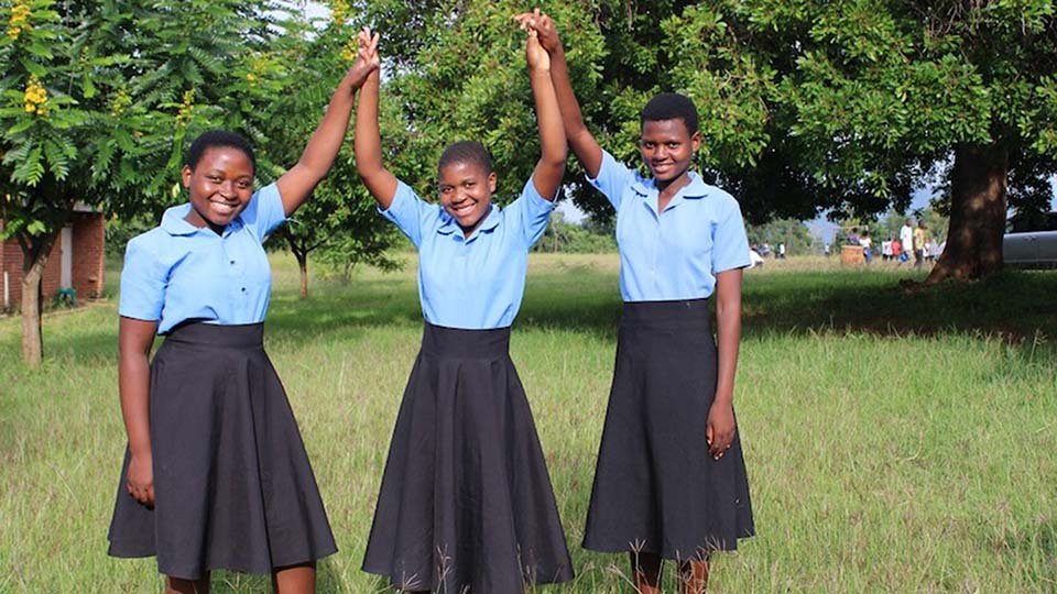 Three girls in matching blue shirts and black skirts hold hands and raise them joyfully on a grassy field, with trees in the background.