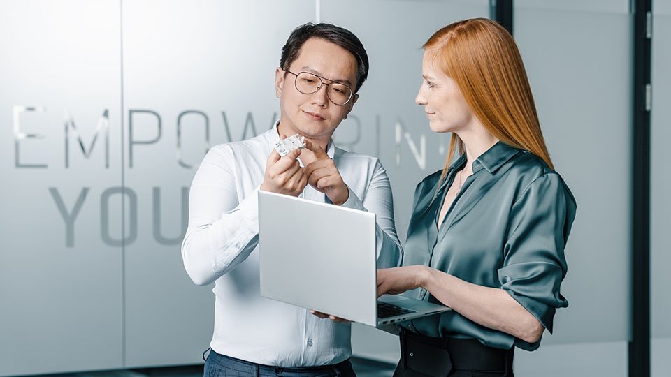 Two colleagues in an office discussing a power module, one holding a laptop. The word "EMPOWERING" is partially visible on a glass wall.