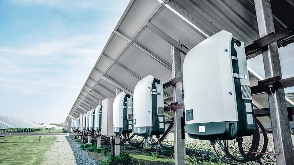 Row of solar inverters under solar panels in a field, converting solar energy into usable electricity, with a clear blue sky above.