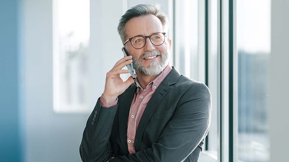 Man with glasses smiling, talking on phone by a window, wearing a suit jacket and pink shirt.