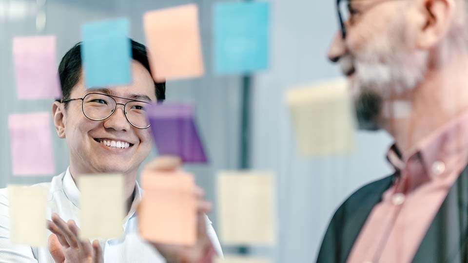 Two colleagues smiling and discussing ideas with colorful sticky notes on a glass board in the foreground.